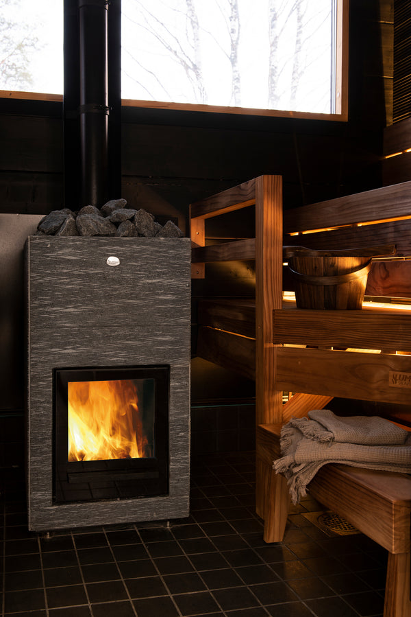 Wood-burning stove in a sauna with wooden benches and a window showing trees.
