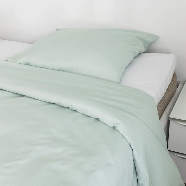 A green SteadyBody duvet cover on a bed, with a pillow in the foreground and nightstand with lamp in the background.
