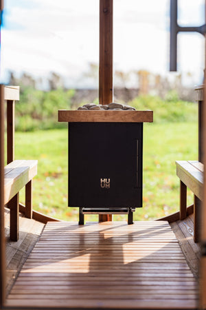Black electric sauna heater with wooden mantel on a wooden deck with a garden view.
