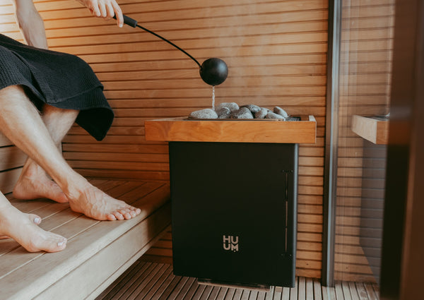 Person sitting in a sauna with a black HUUM Electric sauna heater  on a wooden platform.
