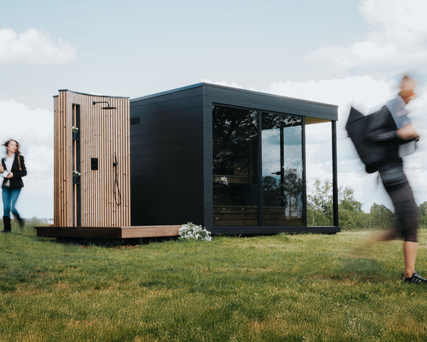 Modern outdoor sauna with glass walls on a grassy area with people walking by.
