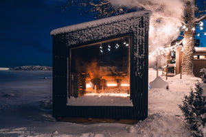 Outdoor fireplace in a snowy landscape with lights and a cabin in the background